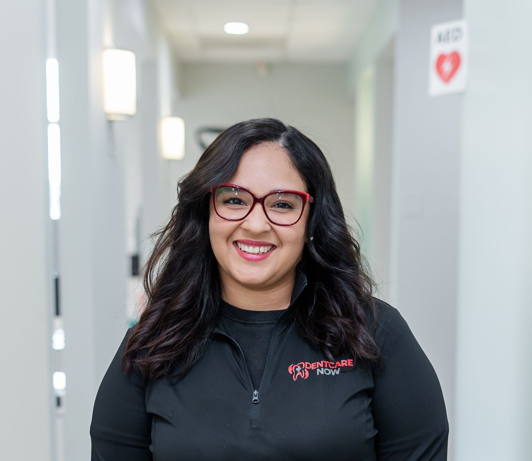 Smiling person in a black quarter-zip shirt standing in a hallway with an AED sign on the wall.