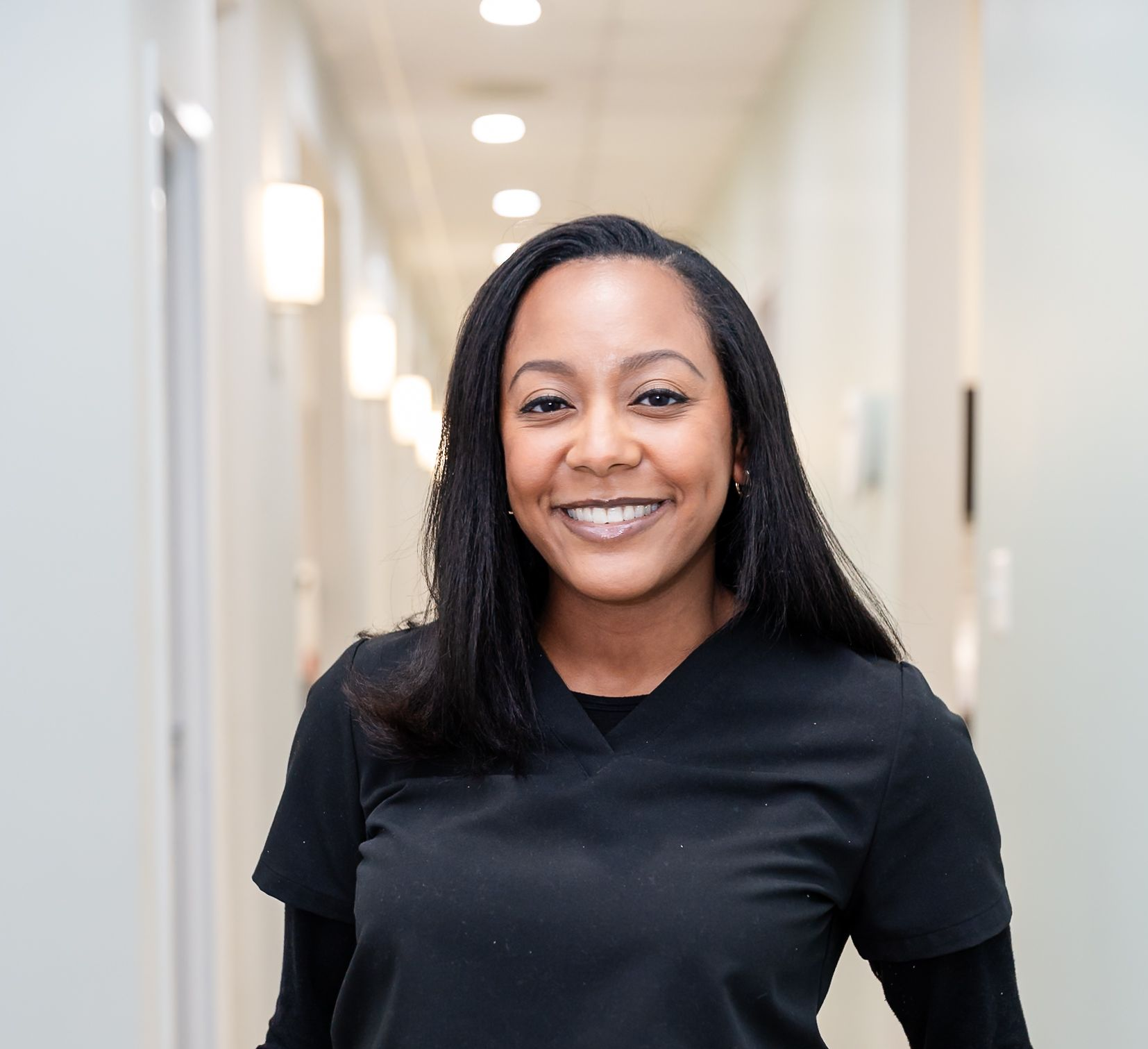 A person wearing black medical scrubs smiling in a brightly lit hallway.