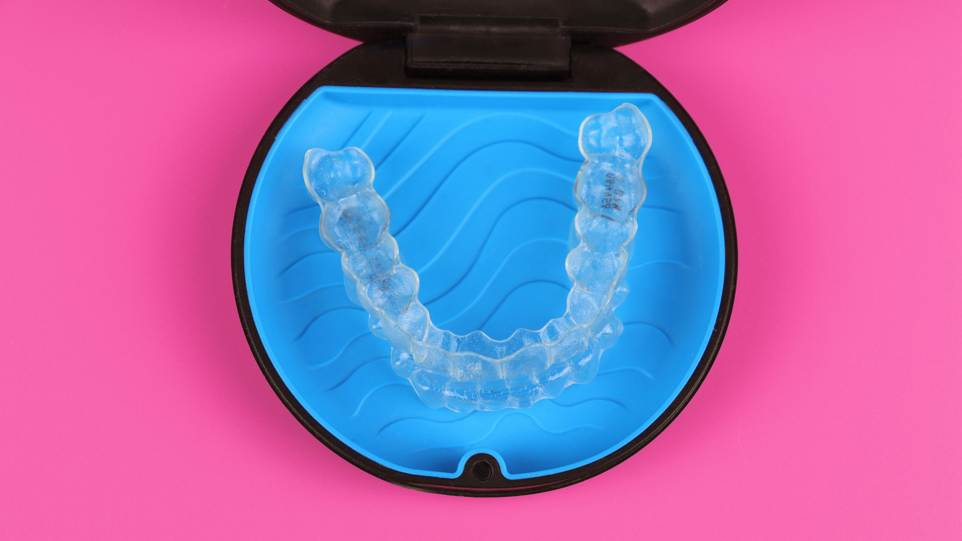 Clear dental aligner in a black case on a bright pink background.