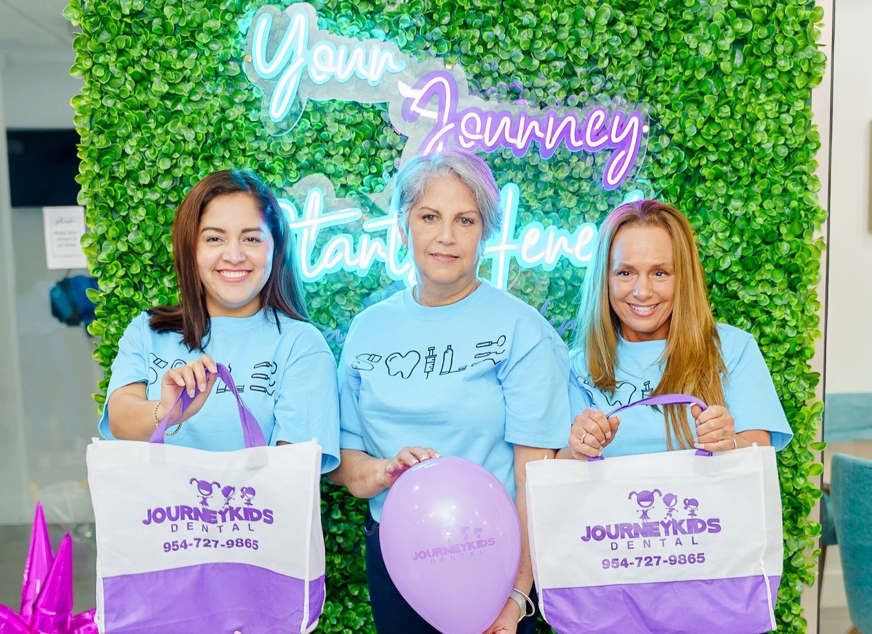 Three women holding purple tote bags and a balloon in front of a green photo wall with neon “Your Journey” sign