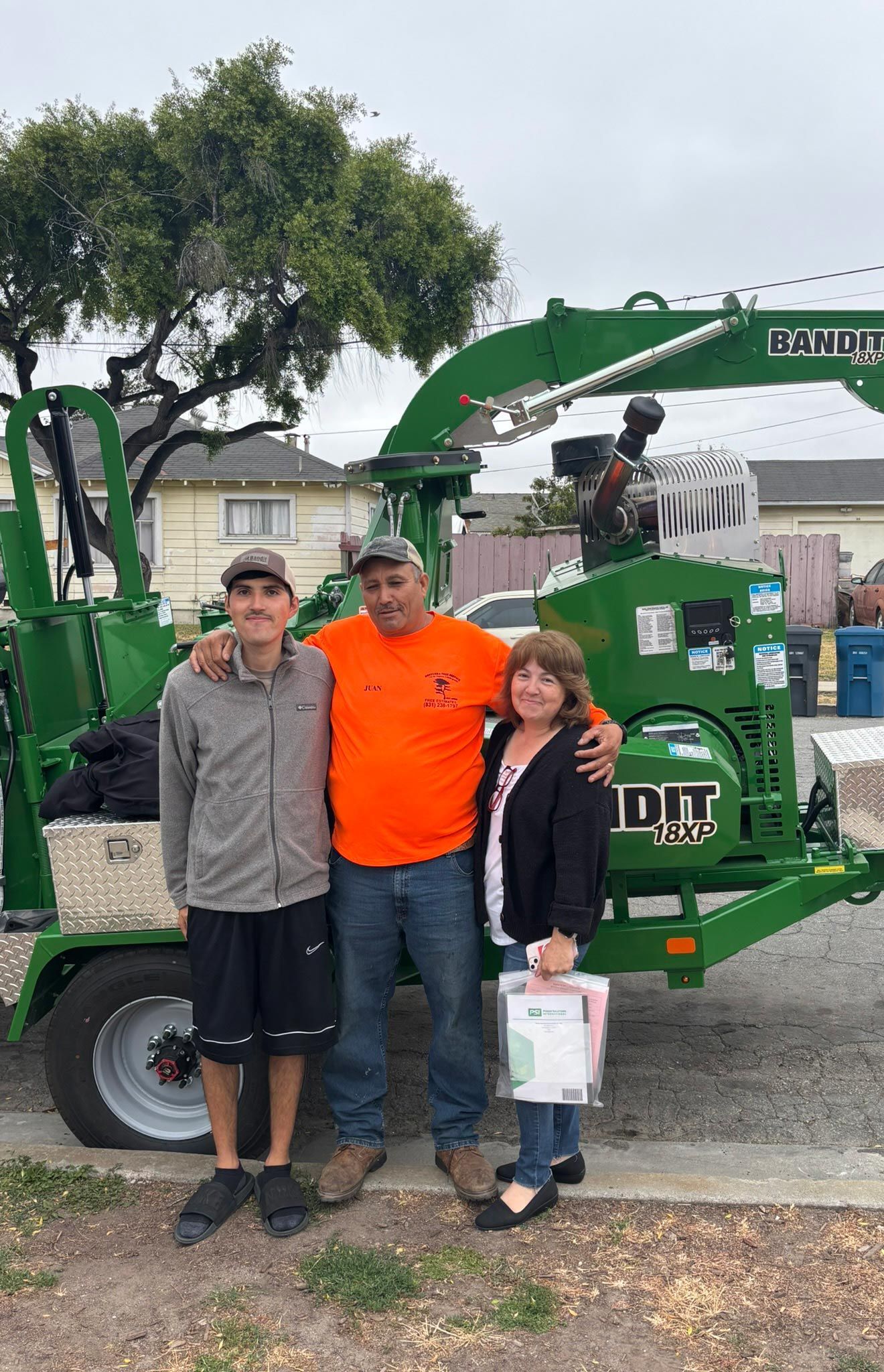 Three people posing by a green wood chipper machine. Man in orange shirt has arms around other two.