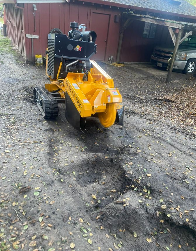 Yellow stump grinder making a trench in muddy ground near a red barn.
