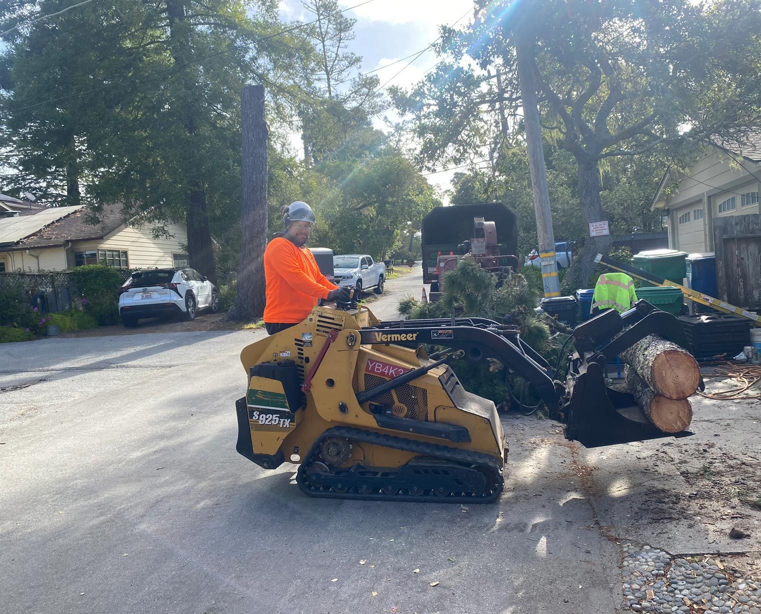 Man operating a small yellow track loader on a street, loading brush and logs into a truck.