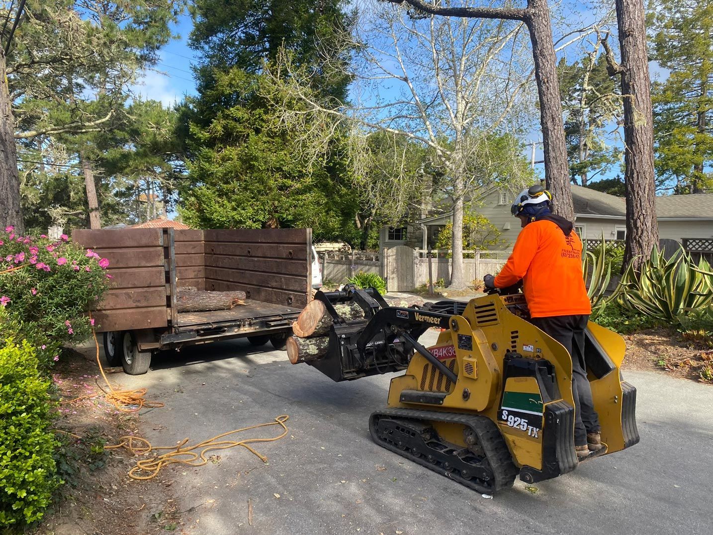 A worker in orange shirt operates a mini-excavator loading wood debris into a trailer on a paved road.
