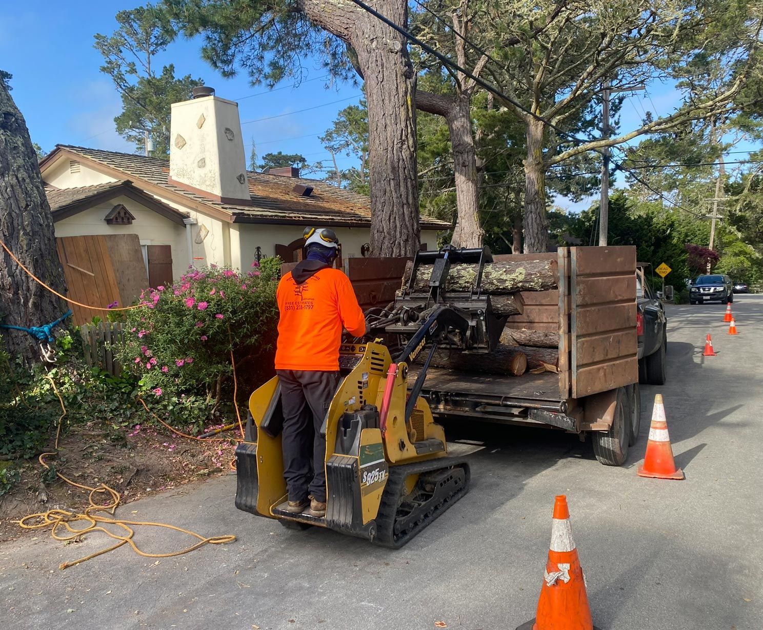 Arborist in orange shirt operating a chipper to load wood debris into a truck, street setting, blue sky.