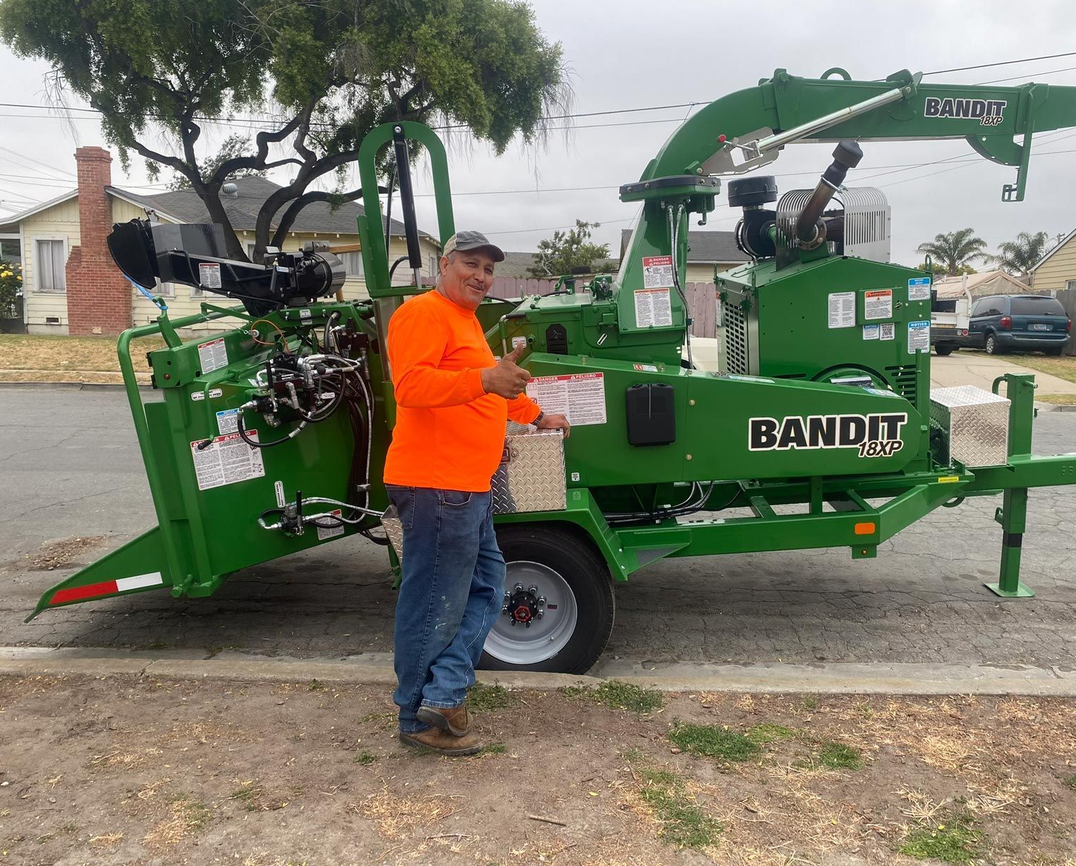 Man in orange shirt gives thumbs-up next to a green Bandit chipper on a street.