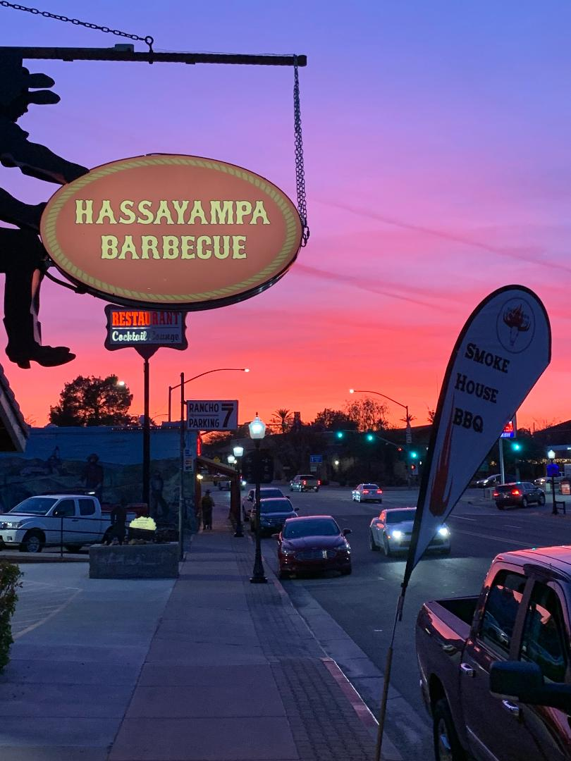 Hassayampa Barbecue sign at sunset on a city sidewalk, with cars and a purple-pink sky