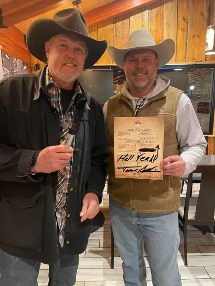 Two cowboy-hatted men posing indoors, one holding a framed award plaque.
