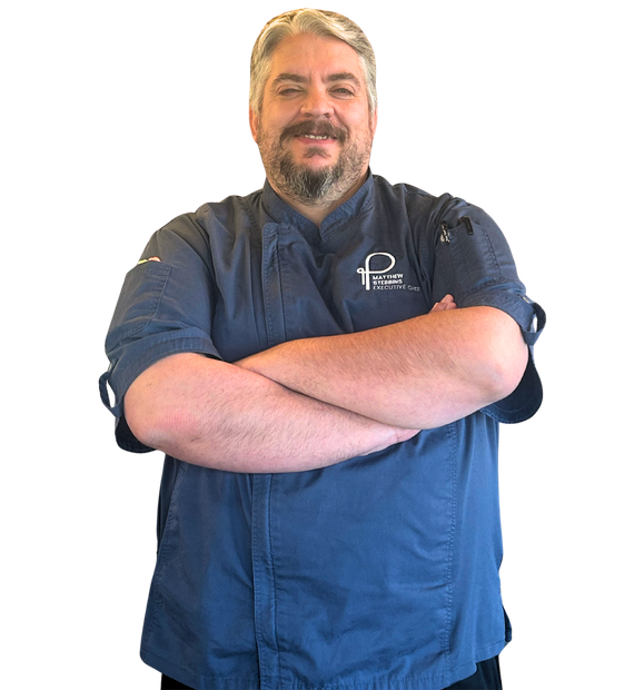 A person with a beard, wearing a blue chef's uniform, stands with arms crossed.