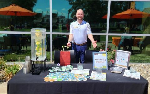 A person stands behind a table with a drink dispenser, a red gift bag, and informational signs.