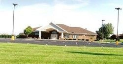 Low brick building with a sloped roof beside a large grassy field and parking lot under a clear sky