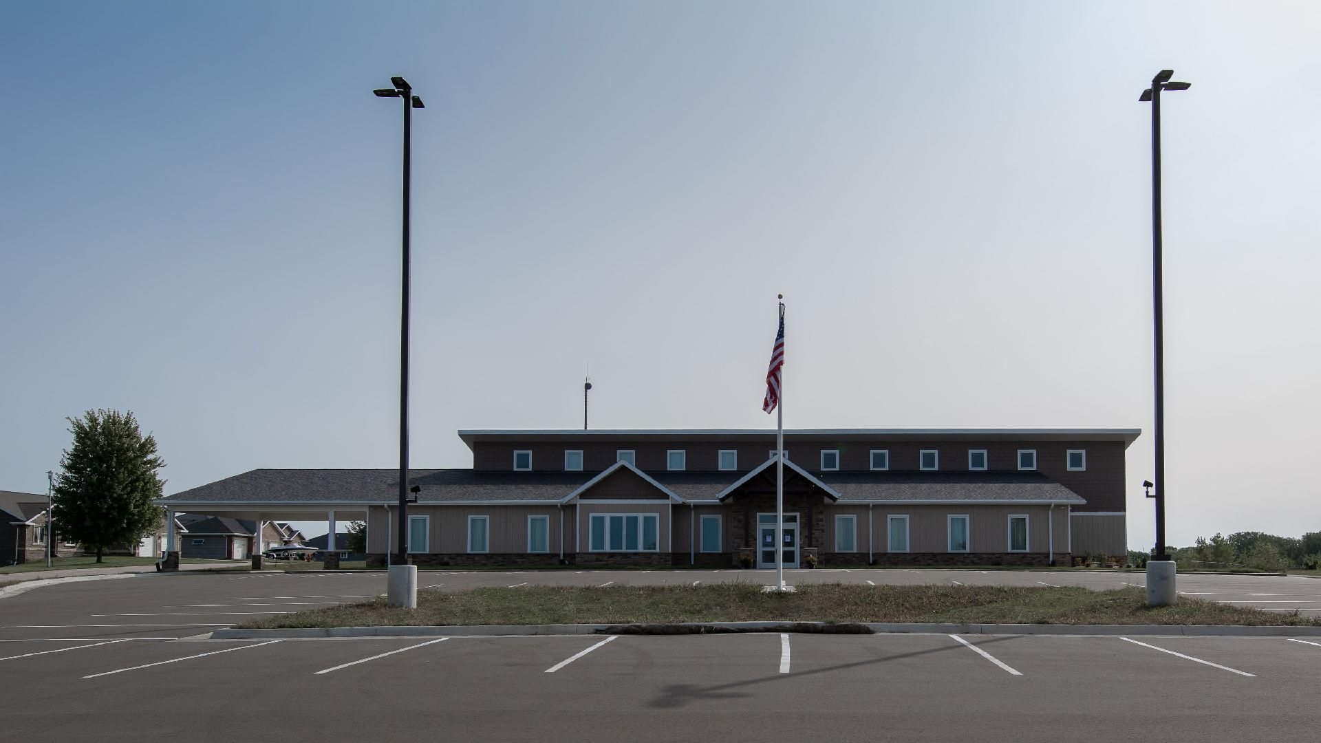 Small airport terminal with an American flag, empty parking lot, and tall light poles under a clear sky.
