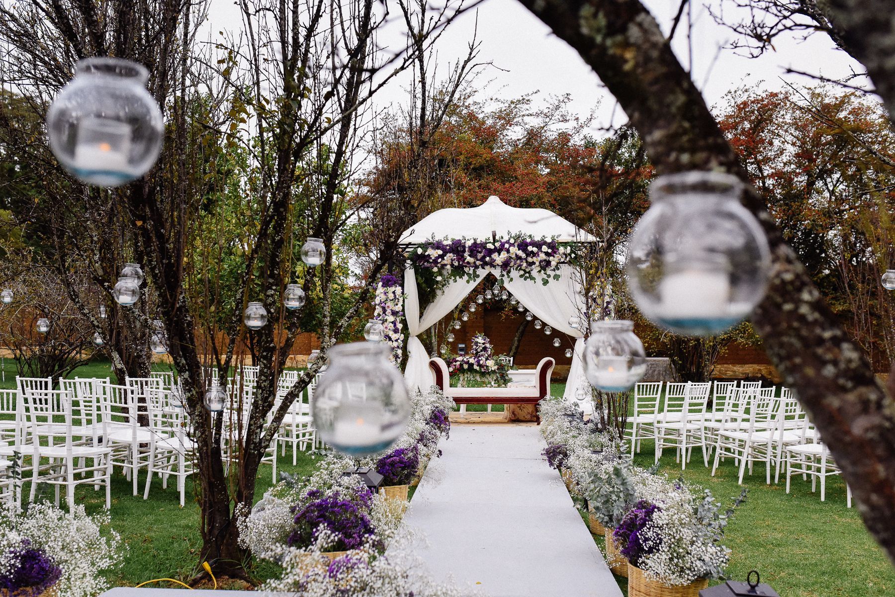 A wedding ceremony is taking place in a park with candles hanging from trees.