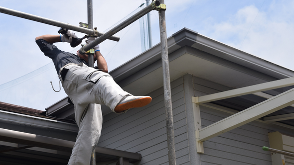 Construction worker on scaffolding, working near a house's edge. Wearing gloves, light pants, and a dark shirt.
