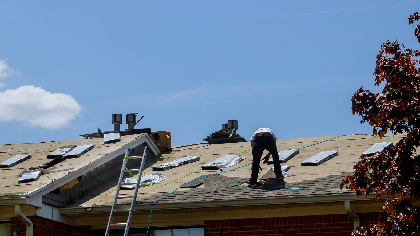 Roofer working on a roof, removing shingles under a blue sky, ladder visible.