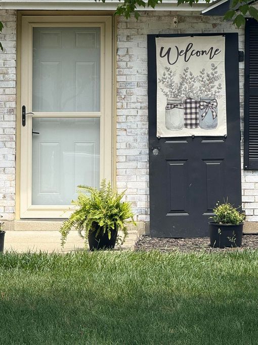 Front porch with a pale door, black door, welcome sign, and potted plants beside a grassy yard.