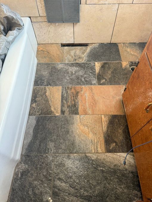 Narrow tiled bathroom floor with mixed gray and tan stone tiles, beside a white tub and wooden cabinet.