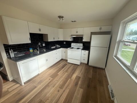 Bright kitchen with white cabinets, black backsplash, wood floors, and a window by the fridge.