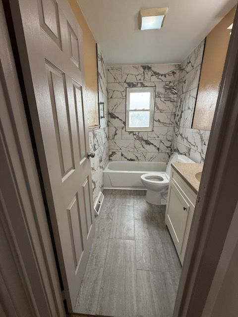 Narrow bathroom with white tub, toilet, vanity, and gray wood-look floor; marble-patterned wall around window.