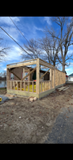 Partially built wooden house frame on a dirt lot under a blue sky with bare trees