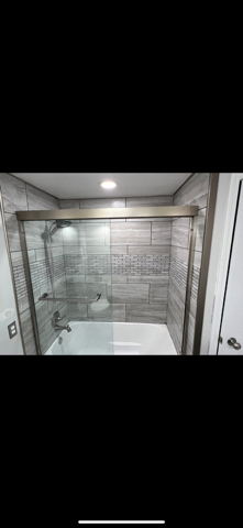 Modern bathroom with glass shower enclosure, gray tile walls, and white bathtub under a ceiling light