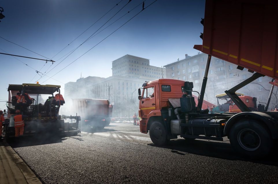 Construction vehicles on a dusty road with a red truck and hazy buildings in the background