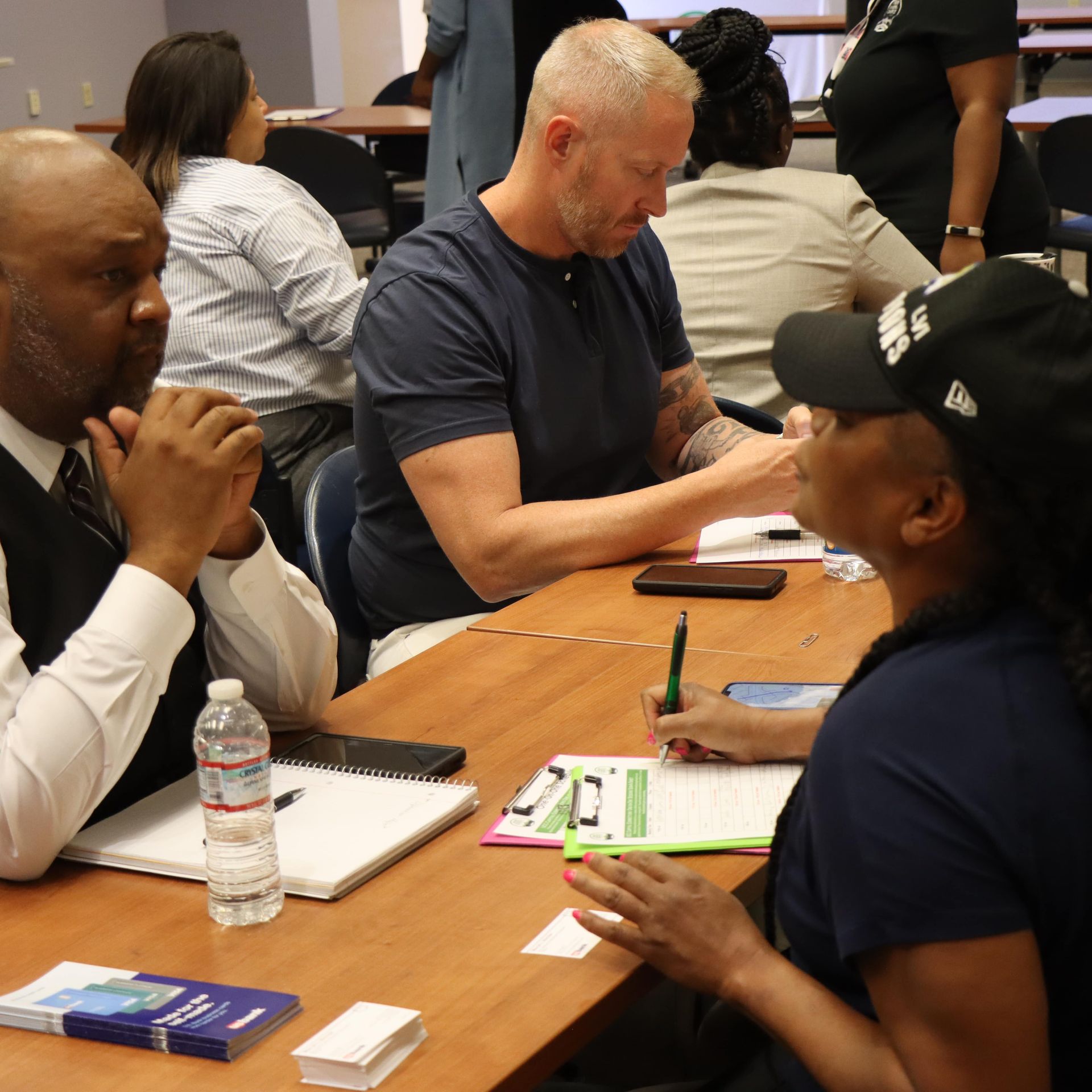 People sitting at a table, taking notes. One man with a beard, another in a suit, and a woman in a hat are visible.