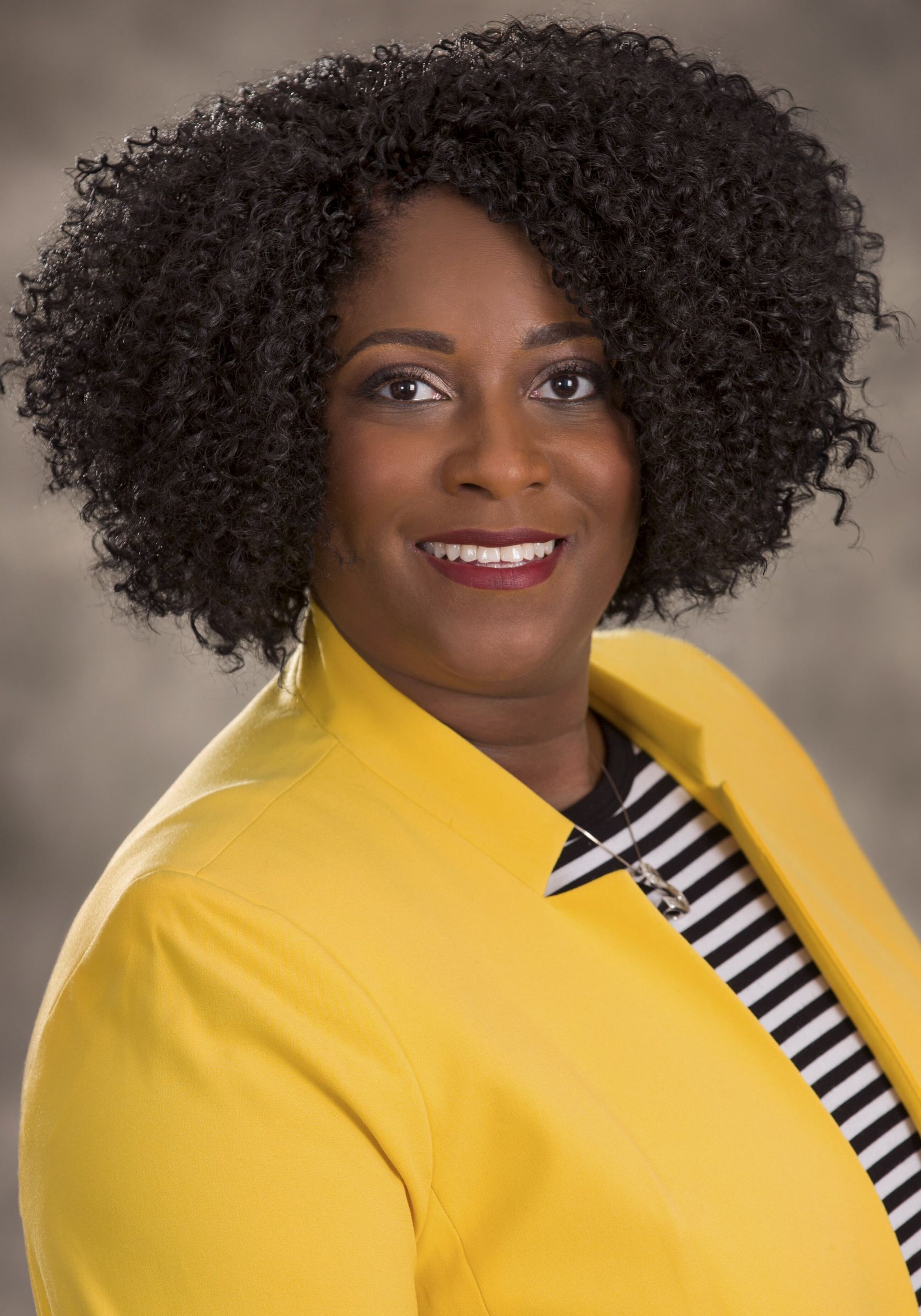 Woman with curly hair in a yellow blazer and striped top smiling.