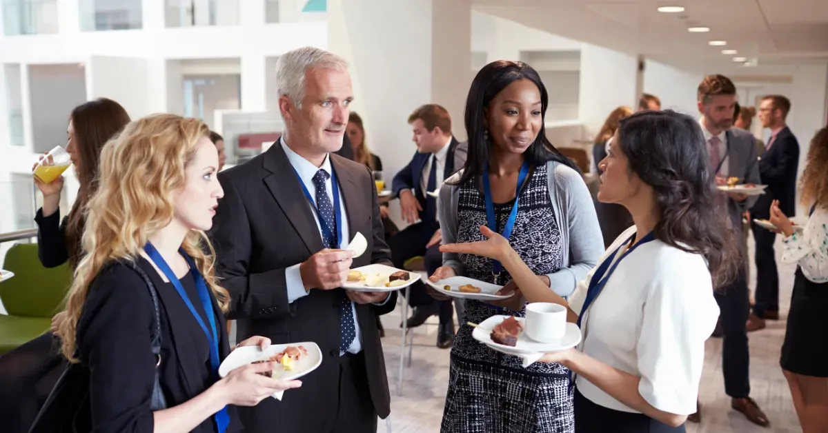 People networking at a conference, some holding food and drinks, in a bright modern space.
