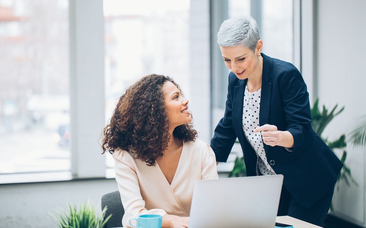 Two women looking at a laptop in a modern office, one pointing, smiling, and explaining to the other.