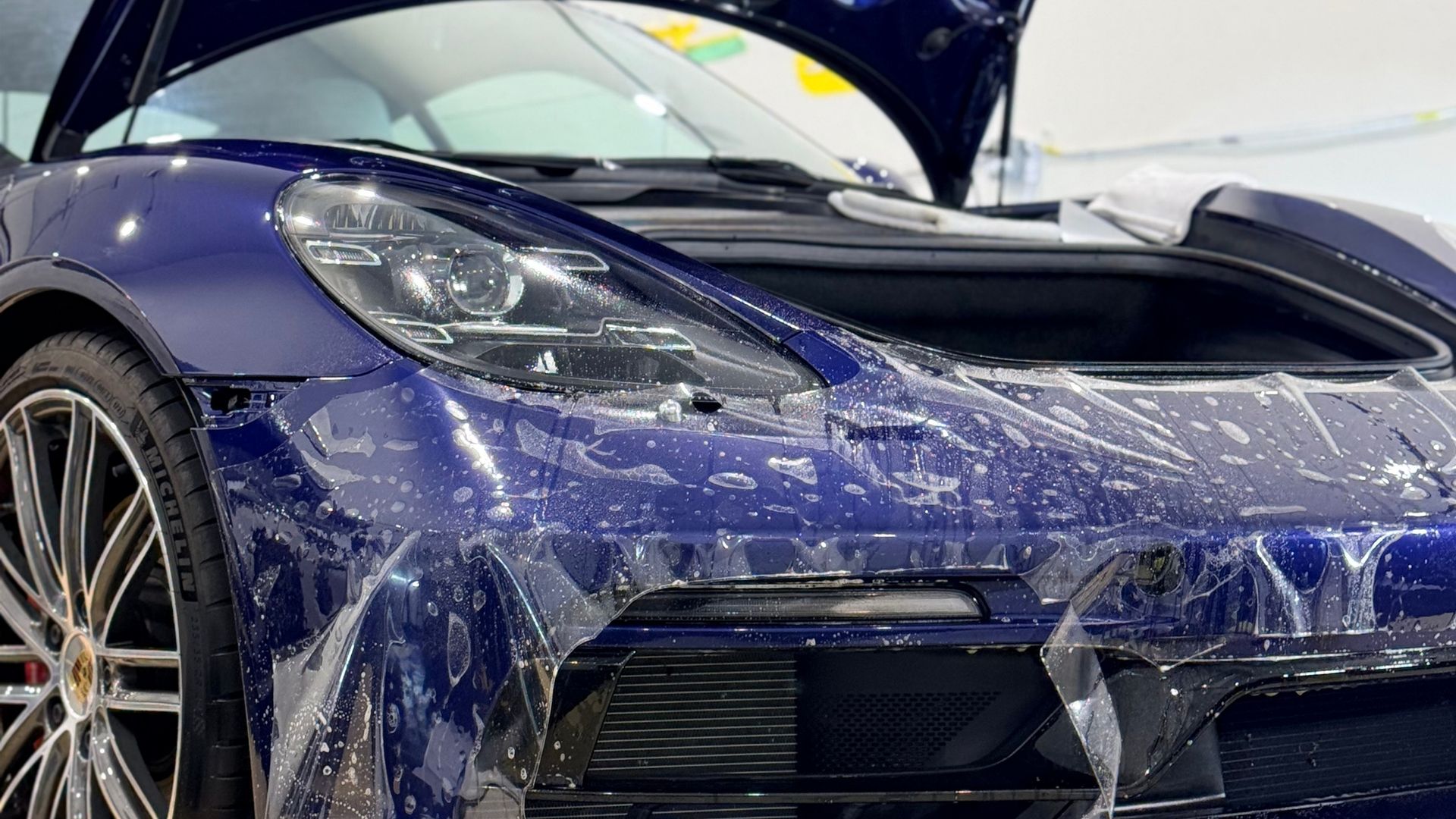 Blue sports car covered in water droplets, front view with hood open in a showroom