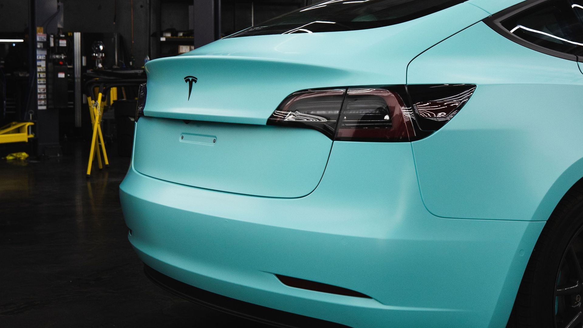 Rear view of a light turquoise Tesla sedan in a garage, showing the trunk and taillights.