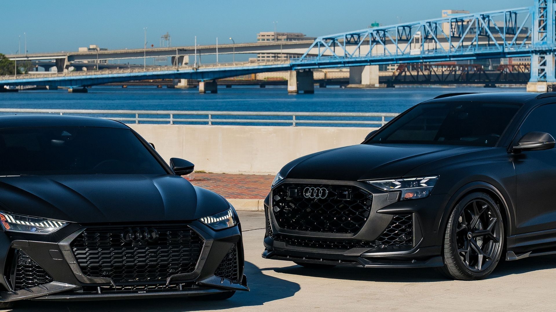 Two black sports cars parked by the water with a bridge in the background
