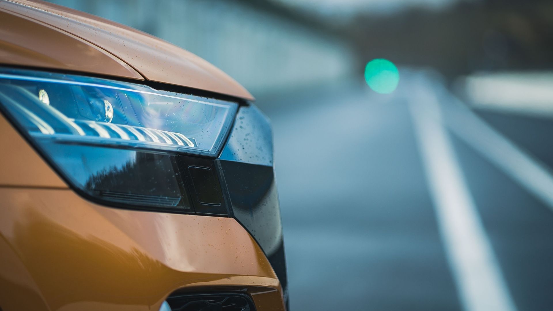 Close-up of a yellow car headlight on a wet road with blurred lane markings and a green light in the distance