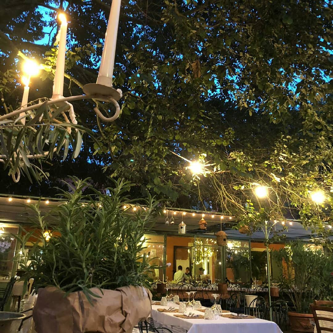 Ristorante all'aperto al tramonto, con tavoli apparecchiati e candele accese sotto gli alberi e le lucine.
