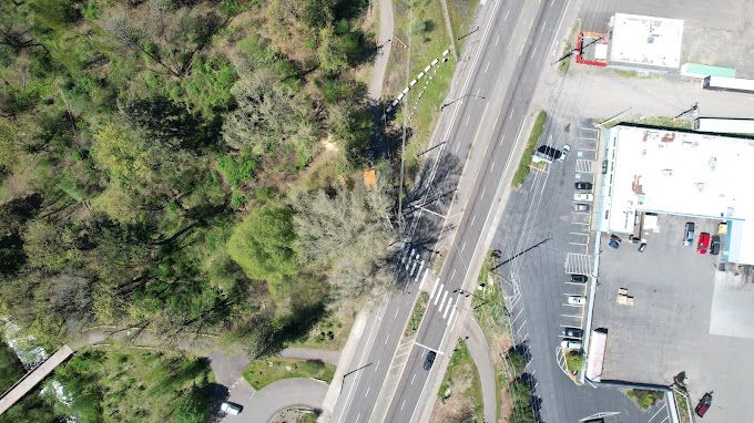 Aerial view of a road with a crosswalk, adjacent to a wooded park area and a commercial building with parking.