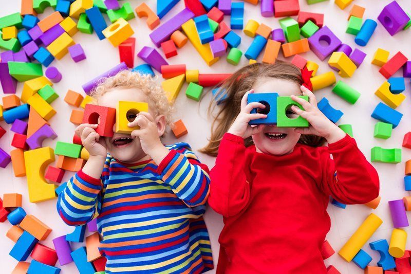 Two children with colorful blocks, holding them up to their eyes, smiling on a white surface scattered with blocks.