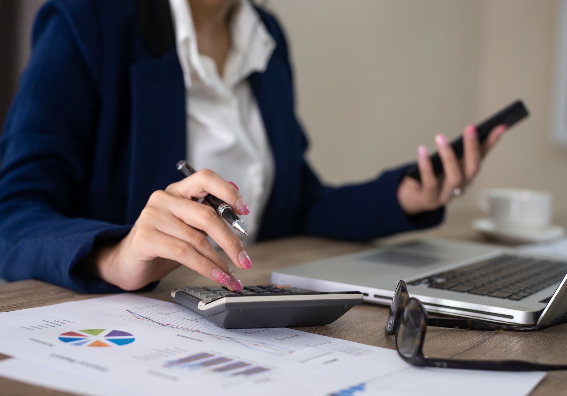 Woman in blazer uses calculator, laptop, and phone, with financial documents.