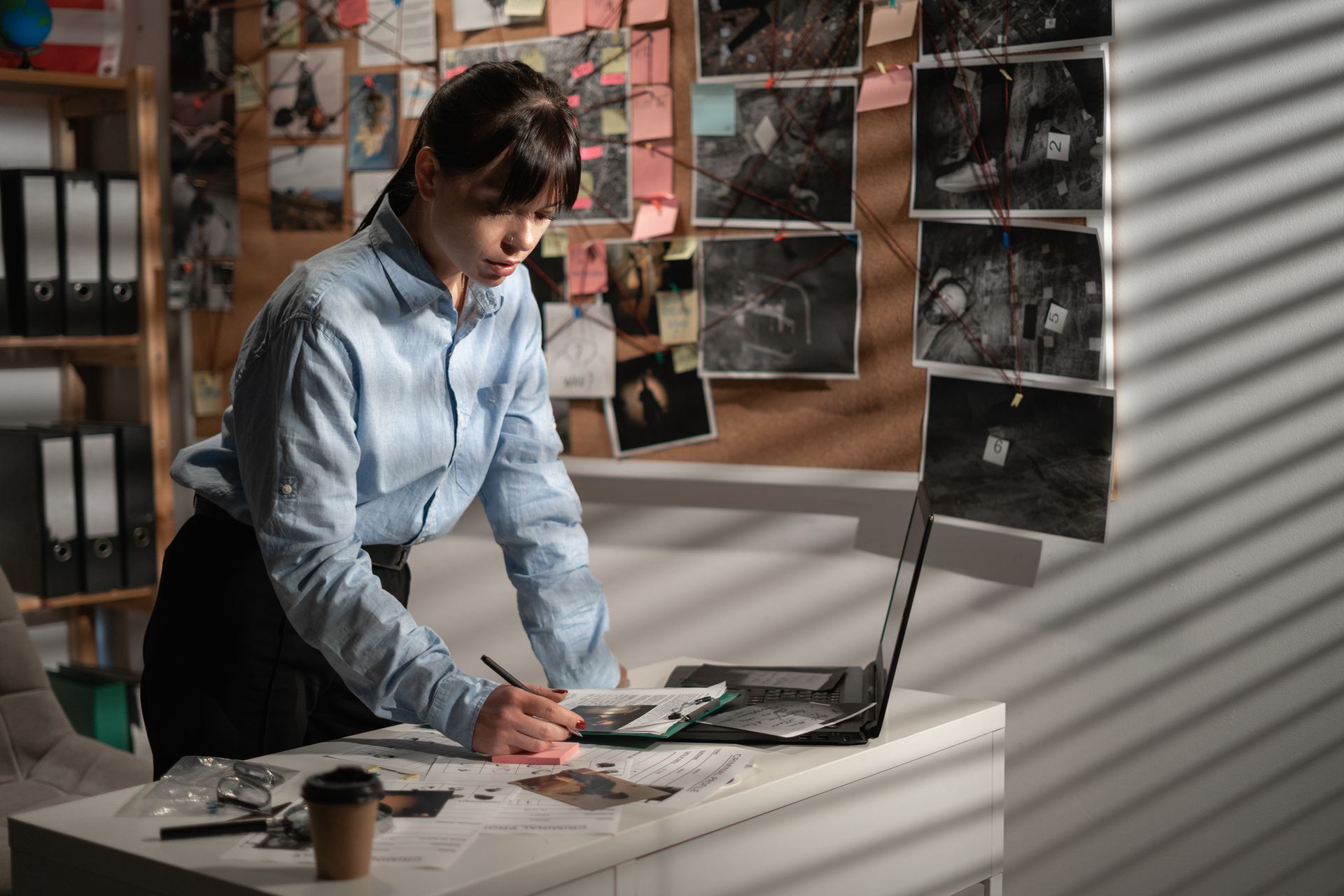 Woman in blue shirt, investigating crime scene photos, working on laptop at desk, in office.
