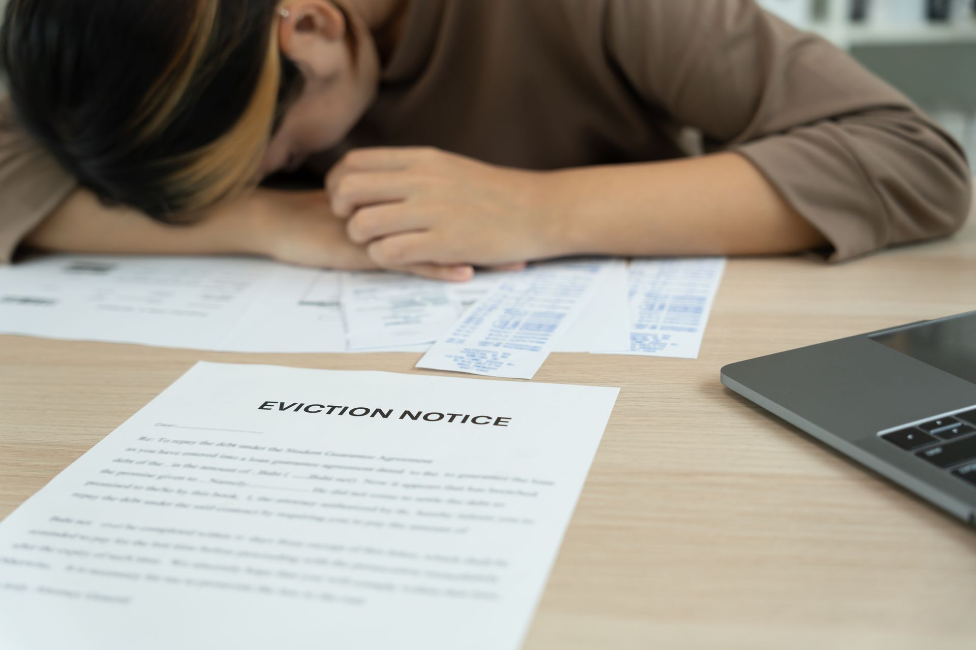 Woman with head on desk, overwhelmed by bills and eviction notice. Laptop nearby.