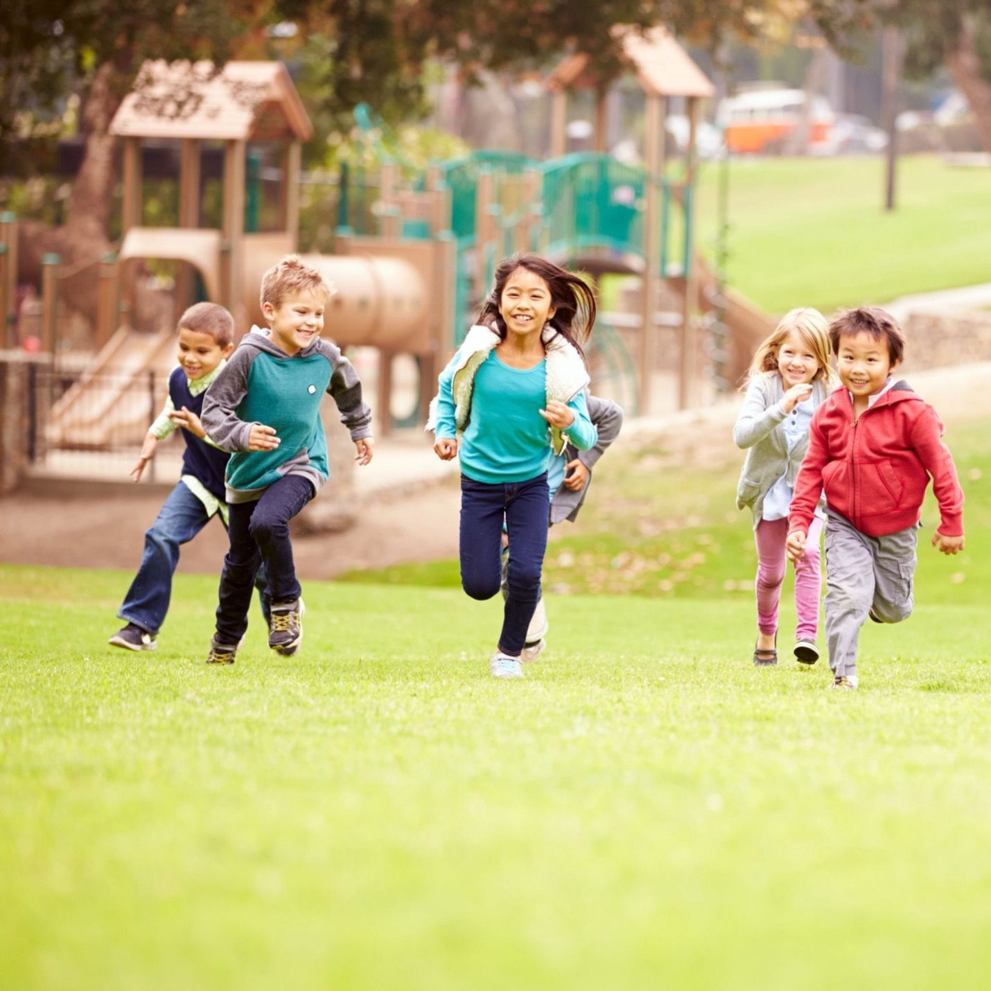 A group of children running in a park with a playground in the background.