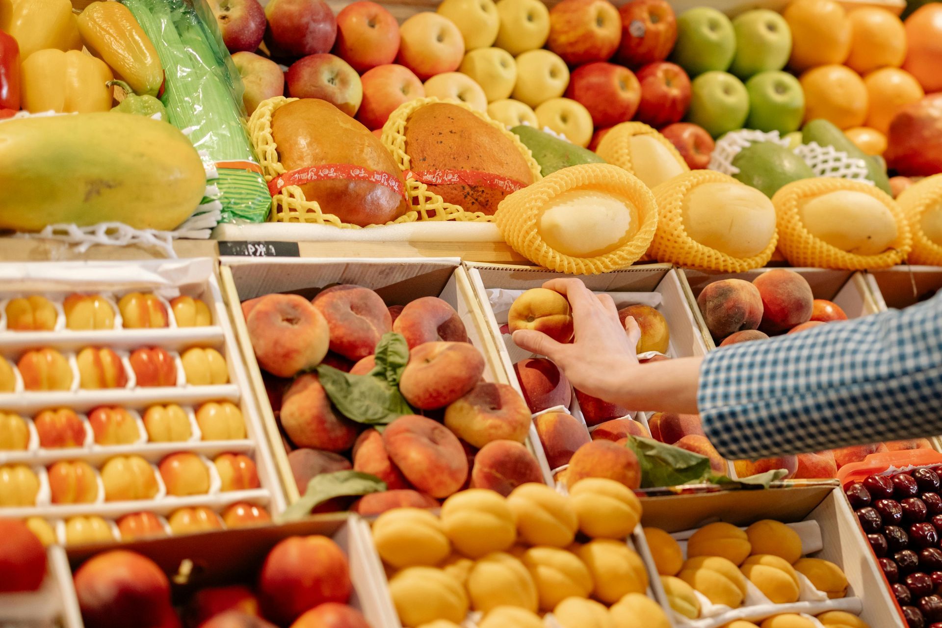 Hands reaching for peaches at a colorful fruit market stall with assorted fresh produce.