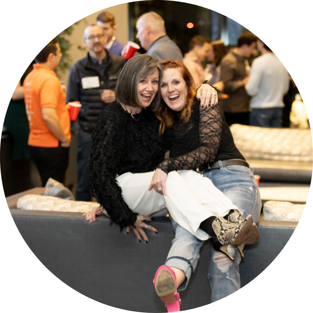 Two women smiling, arm in arm, sitting on a couch at a social gathering.