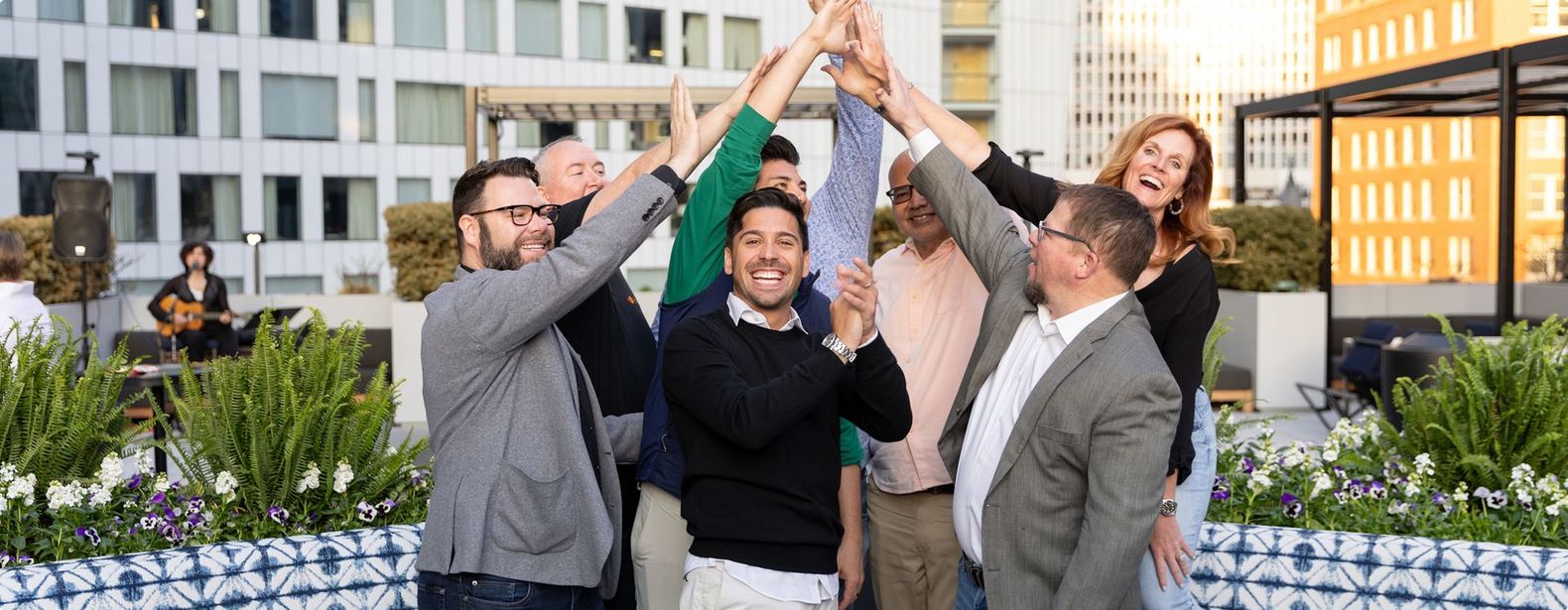 Group of people celebrating outdoors with hands raised. Buildings in the background.