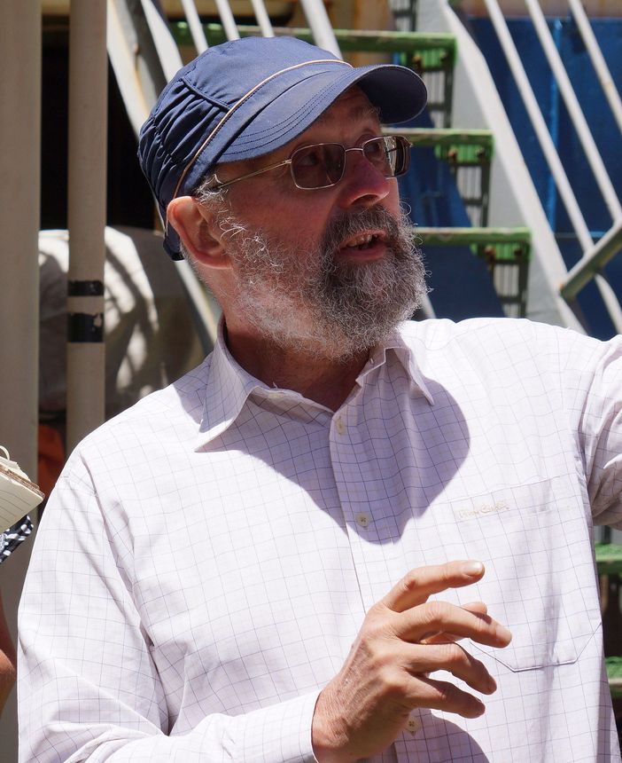 Man with a beard, wearing a denim cap and glasses, gesturing. Standing outdoors, with stairs in the background.
