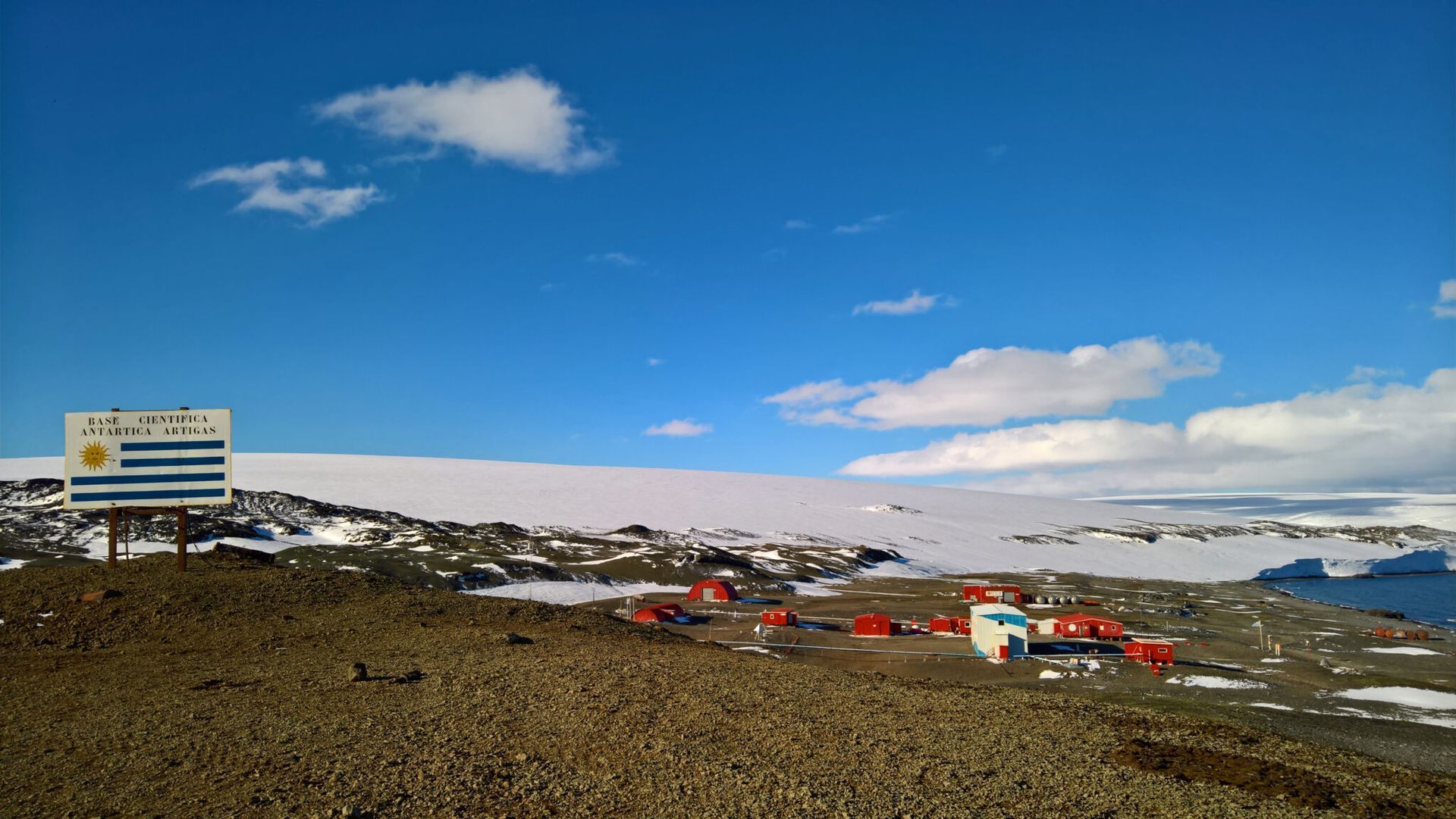 Uruguayan Antarctic base with flag, red buildings, and snowy landscape under a blue sky.