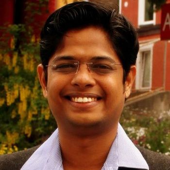Man with glasses smiling outdoors in front of a building and greenery.