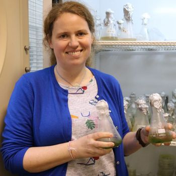 Woman in blue cardigan smiles, holding green liquid flasks in a lab setting, with more flasks in the background.