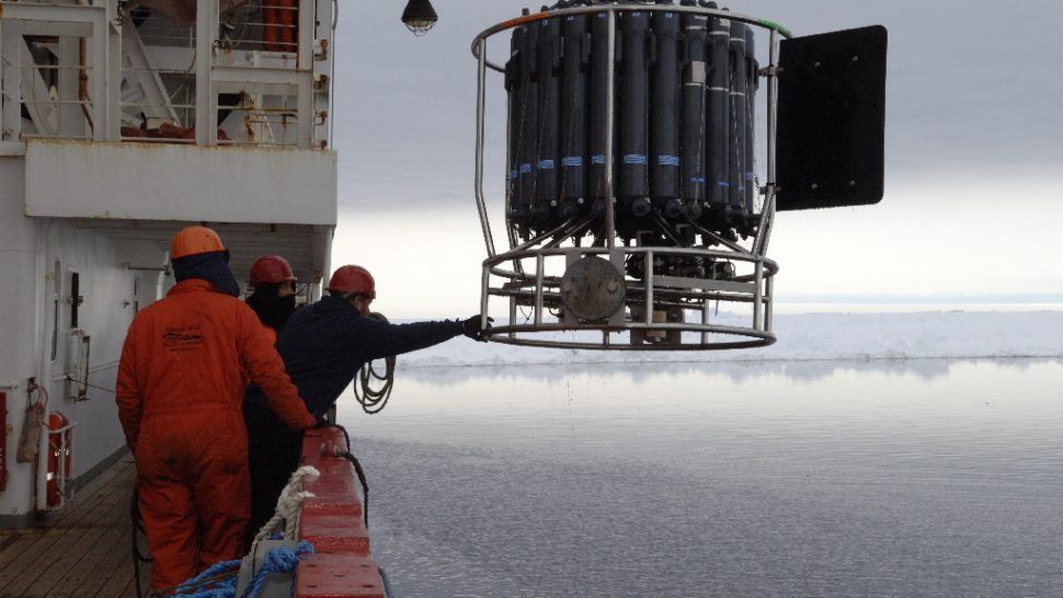 Men on ship deck lower a large cylindrical instrument into icy water.