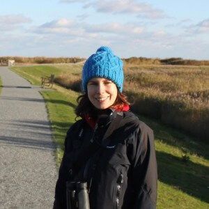 Woman in blue hat and black jacket smiles outdoors, holding binoculars; path and marsh in background.